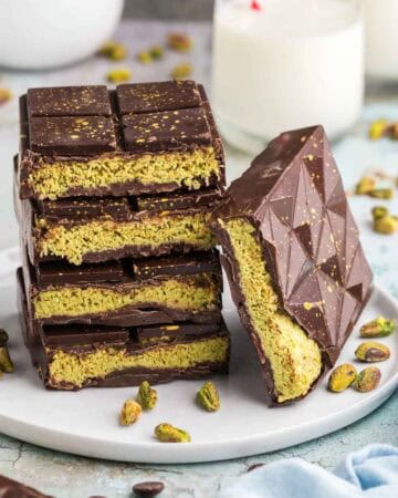 Side shot of four cut open Dubai chocolate bars stacked on top of each other. A fifth bar is laying on its side leaning up against the stack. It too is cut open to reveal a light green filling of pistachio cream and toasted kataifi. In the background is a bowl of green pistachios and a glass of milk. There are also pistachios scattered around the scene.