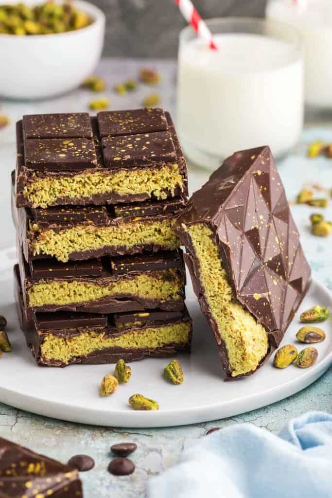 Side shot of four cut open Dubai chocolate bars stacked on top of each other.  A fifth bar is laying on its side leaning up against the stack.  It too is cut open to reveal a light green filling of pistachio cream and toasted kataifi.  In the background is a bowl of green pistachios and a glass of milk.  There are also pistachios scattered around the scene.