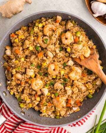 Overhead shot of a grey fry pan filled with Shrimp and Chicken Fried Rice. The rice is a golden orange brown color, and is dotted with plenty of orange carrots, and green celery. The shrimp are big and pink layered throughout the dish. Fresh onion, garlic, ginger and green onion are laying around the frypan.