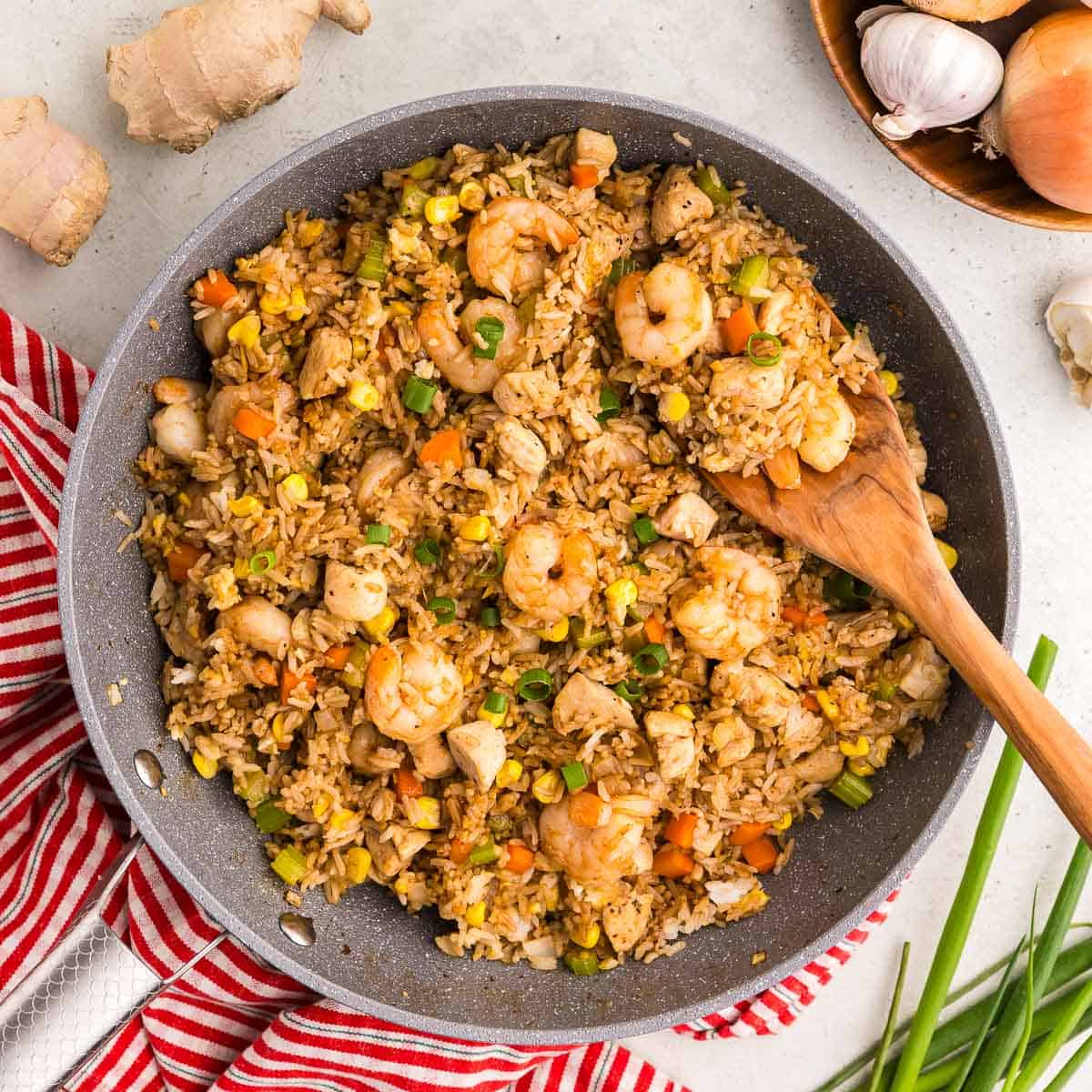 Overhead shot of a grey fry pan filled with Shrimp and Chicken Fried Rice. The rice is a golden orange brown color, and is dotted with plenty of orange carrots, and green celery. The shrimp are big and pink layered throughout the dish. Fresh onion, garlic, ginger and green onion are laying around the frypan.