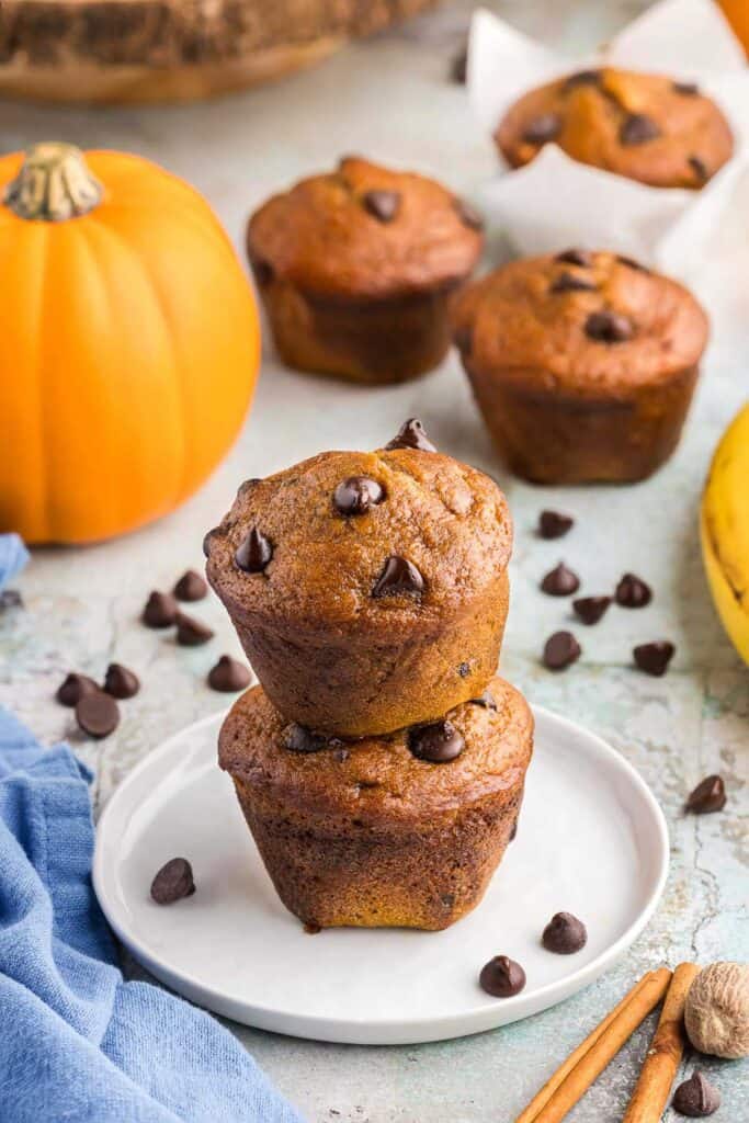 Side shot of a 2-stack of pumpkin banana bread muffins. The top muffin has four melty chocolate chips sitting on top. The stack sits on top of a small white plate with a few chocolate chips. In the foreground on the right are a couple of cinnamon sticks and a nutmeg. On the left is a blue linen. In the background is a pumpkin and some more muffins.