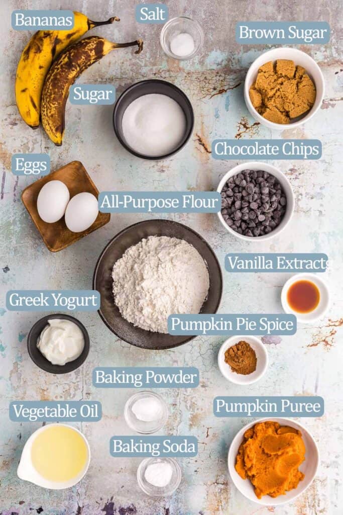 An overhead shot of all the ingredients needed to make pumpkin and banana muffins. Ingredients are: bananas, salt, sugar, brown sugar, chocolate chips (optional), eggs, all-purpose flour, vanilla extract, pumpkin pie spice, Greek yogurt, baking powder, baking soda, vegetable oil, and pumpkin puree.