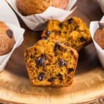 Close up side shot of a wooden cake platter with Pumpkin Banana Bread Muffins on it. Most are in white tulip muffin liners but the one in the middle has no liner and has been torn open to reveal a moist orangey colored muffin with lots of melty chocolate chips inside.