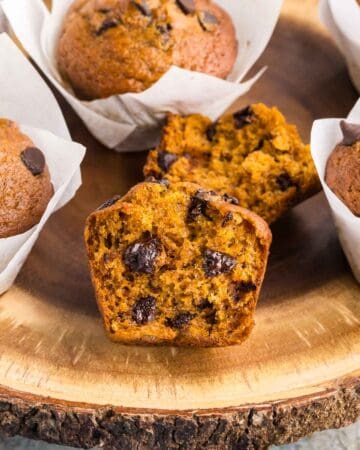 Close up side shot of a wooden cake platter with Pumpkin Banana Bread Muffins on it. Most are in white tulip muffin liners but the one in the middle has no liner and has been torn open to reveal a moist orangey colored muffin with lots of melty chocolate chips inside.