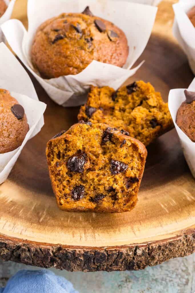 Close up side shot of a wooden cake platter with Pumpkin Banana Bread Muffins on it. Most are in white tulip muffin liners but the one in the middle has no liner and has been torn open to reveal a moist orangey colored muffin with lots of melty chocolate chips inside.