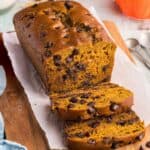A side shot of a loaf of pumpkin chocolate chip bread being sliced open. There are two slices that have been cut and are laying in front of the loaf. The bread part is an orangey/yellow/brown color and there are lots of chocolate chips distributed throughout. In the background a couple of pumpkin decorations are present.
