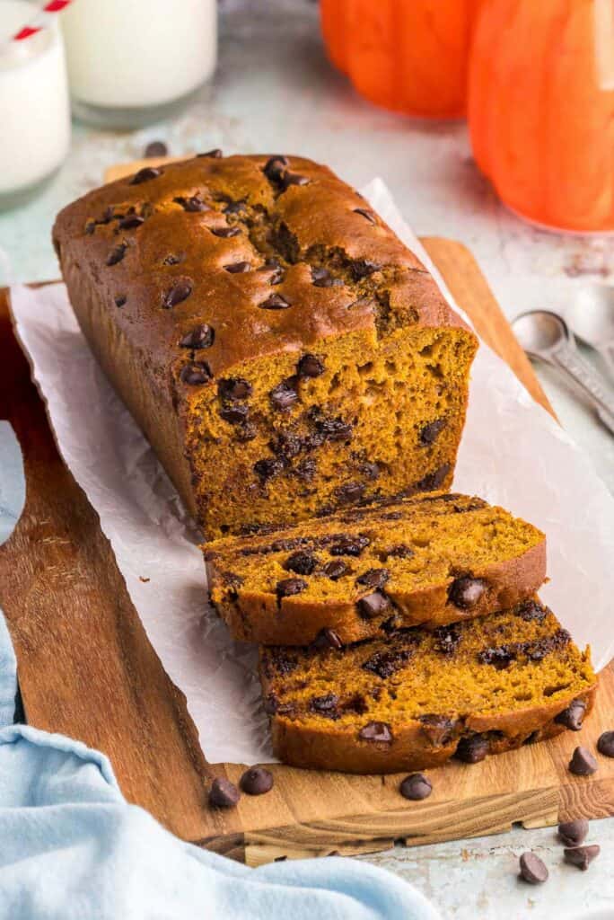 A side shot of a loaf of pumpkin chocolate chip bread being sliced open.  There are two slices that have been cut and are laying in front of the loaf.  The bread part is an orangey/yellow/brown color and there are lots of chocolate chips distributed throughout.  In the background a couple of pumpkin decorations are present.