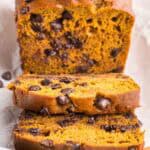 Front view of a loaf of pumpkin chocolate chip bread sliced open. Two slices have fallen forward and there tops are easily seen. The rest of the loaf is in the background. The loaf is a brown/orangey color with lots of melty chocolate chips present.