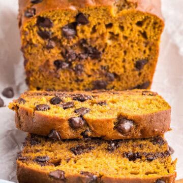 Front view of a loaf of pumpkin chocolate chip bread sliced open. Two slices have fallen forward and there tops are easily seen. The rest of the loaf is in the background. The loaf is a brown/orangey color with lots of melty chocolate chips present.