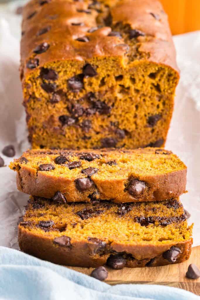 Front view of a loaf of pumpkin chocolate chip bread sliced open.  Two slices have fallen forward and there tops are easily seen.  The rest of the loaf is in the background.  The loaf is a brown/orangey color with lots of melty chocolate chips present.