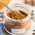 Side shot close up of a glass spice jar filled with an orangey/brown spice with a white label on the front which says "Pumpkin Spice". A silver spoon is resting in the spice jar. In the background are some pumpkins, nutmegs, and whole cloves, as well as a blue linen.