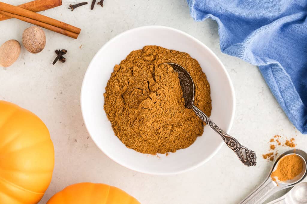 Overhead shot of a small white bowl filled with an orangey/brown spice mixture called Pumpkin Spice.  There is a small metal spoon in the bowl.  The top left corner of the picture has some cinnamon sticks, nutmegs, and cloves peeking in.  The top right corner has a blue linen.  The bottom left corner has the edges of two pumpkins, and the lower right are some measuring spoons.