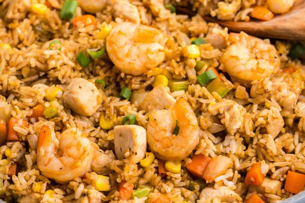 Close up 45 degree angle shot looking into the frypan of fully cooked Shrimp and chicken fried rice. Large shrimp and chunks of chicken are studded throughout the brown-tinted rice and veggies.