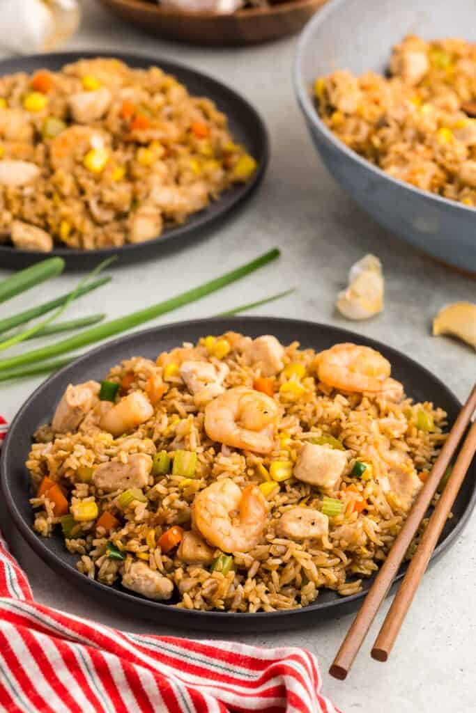 Side shot of a dark grey plate filled with Shrimp and chicken fried rice. There a lots of plump juicy shrimp as well as tender chunks of chicken studded throughout the rice and vegetables. A pair of wooden chopsticks sit on the right hand side of the plate. In the background is another plate filled with fried rice as well as the frypan that served the fried rice.