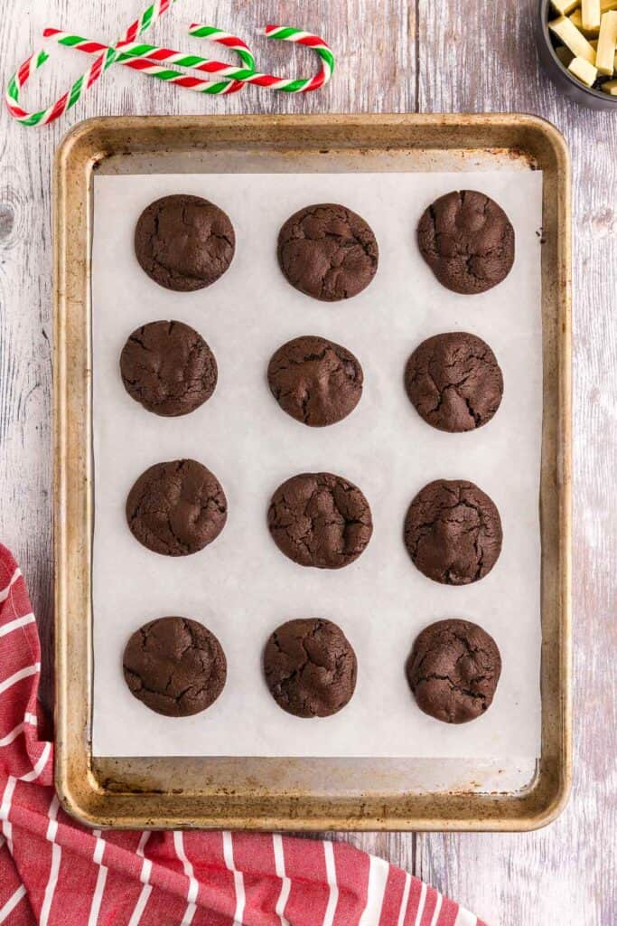 Overhead shot of a baking pan lined with white parchment paper and 12 chocolate peppermint cookies that have just come out of the oven. The cookies are very round and dark brown with a crinkly top.
