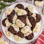 Overhead shot of a large white serving plate filled with chocolate peppermint cookies. In the upper right and lower left corner, more cookies are peaking in to the scene. A red linen is in the bottom right corner, and some cedar boughs and Christmas balls are in the top left corner.