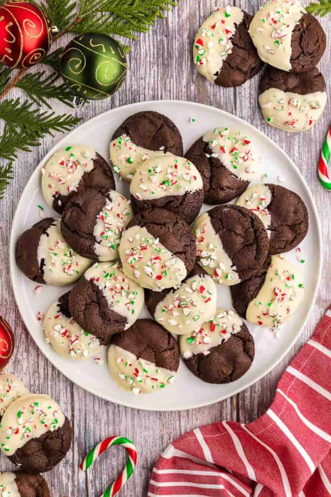 Overhead shot of a large white serving plate filled with chocolate peppermint cookies. In the upper right and lower left corner, more cookies are peaking in to the scene. A red linen is in the bottom right corner, and some cedar boughs and Christmas balls are in the top left corner.