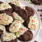 Side shot of a white serving plate filled with Chocolate Peppermint Cookies. Cookies are dark brown and one half of each cookie has been dipped in white chocolate and sprinkled with crushed candy cane.