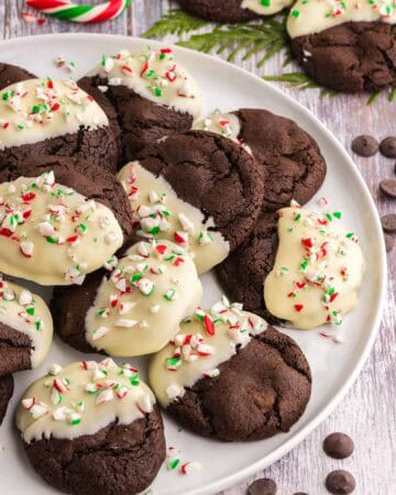 Side shot of a white serving plate filled with Chocolate Peppermint Cookies. Cookies are dark brown and one half of each cookie has been dipped in white chocolate and sprinkled with crushed candy cane.