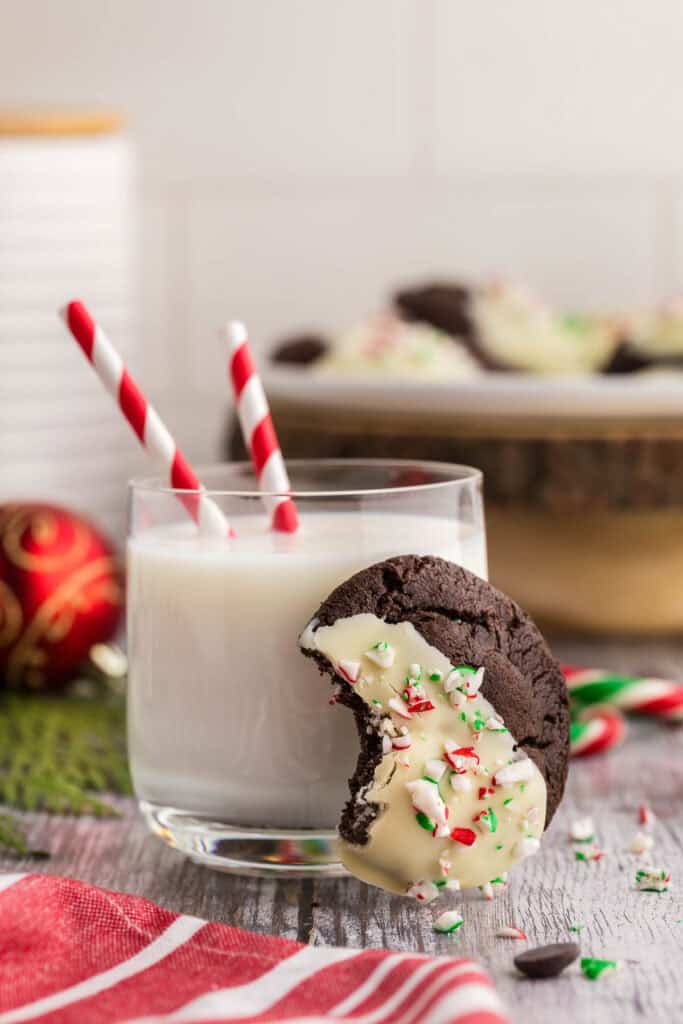Side shot of a chocolate peppermint cookie half dipped in white chocolate and sprinkled with crushed candy cane and a bite taken out of it, is leaning up against a glass of milk.
