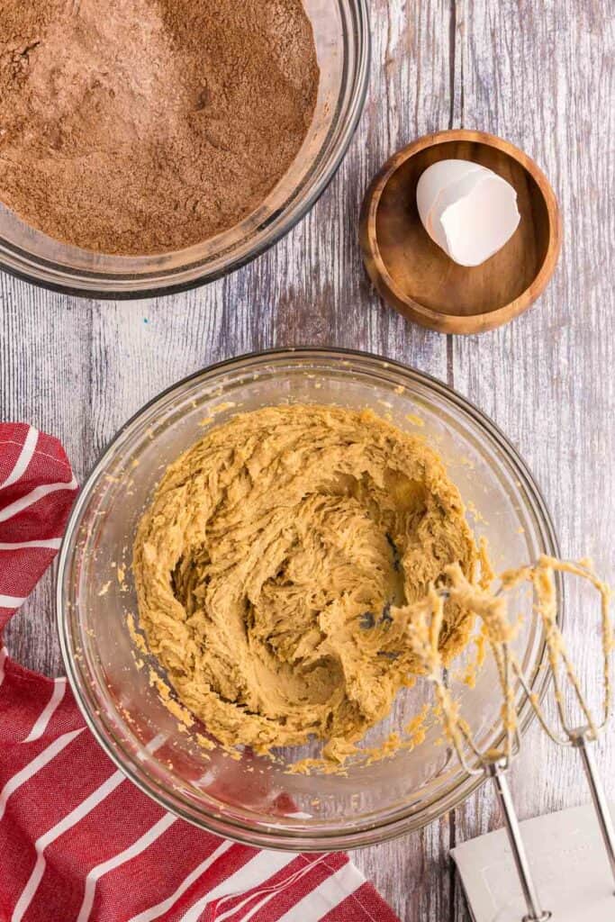 Overhead shot of a glass mixing bowl in the process of making chocolate peppermint cookies. In the mixing bowl the butter and sugars have been creamed together and now the egg and vanilla extract have been incorporated. The mixture is a beigey brown color and is slightly fluffy.