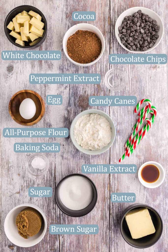 Overhead shot of the ingredients needed to make Chocolate Peppermint Cookies. Ingredients include: All-purpose Flour, white sugar, brown sugar, natural cocoa, white chocolate, semi-sweet chocolate chips, egg, baking soda, peppermint extract, vanilla extract, salted butter, candy canes.