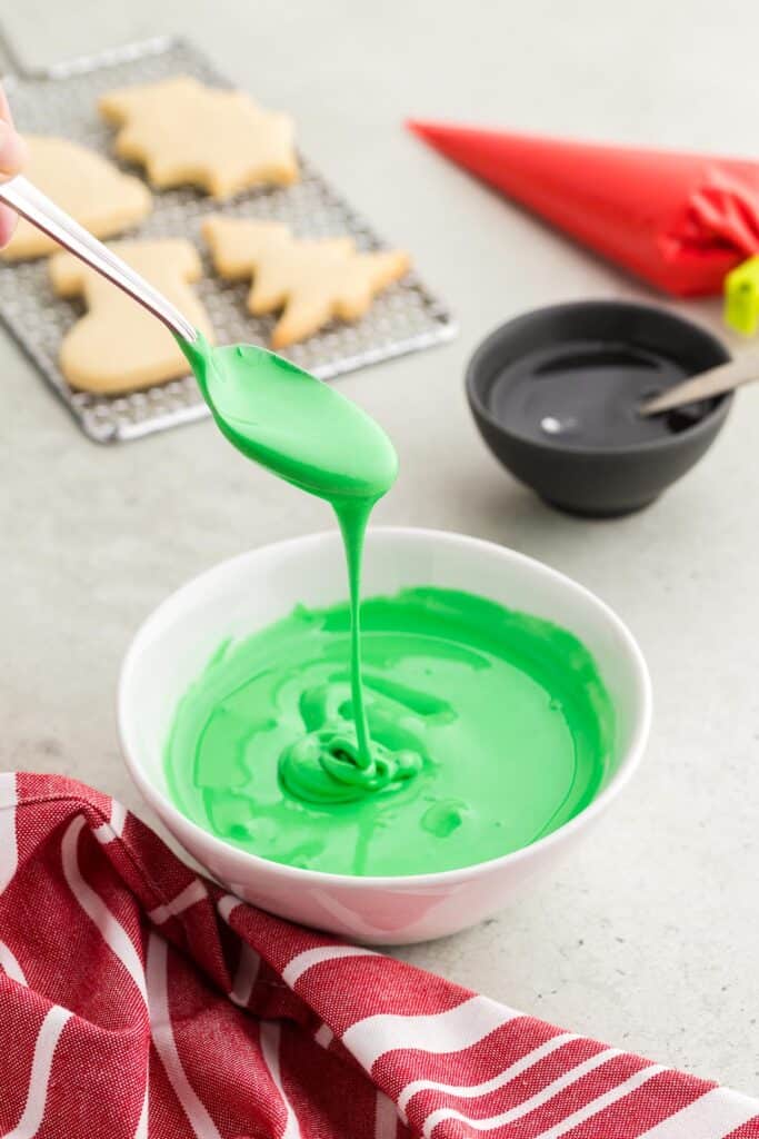 A side shot of green royal icing with meringue powder.  A spoon is drizzling the icing back into the bowl where the consistency is shown as lines that haven't melted back into the rest of the icing yet.  In the background are some sugar cookies waiting to be iced.