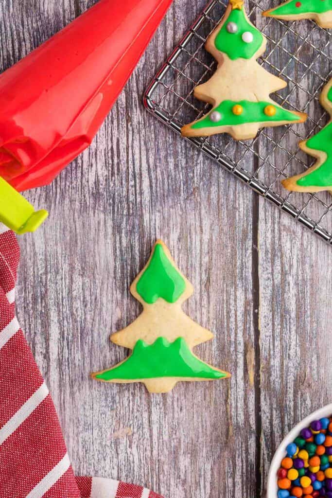Overhead shot of a sugar cookie shaped like a Christmas tree.  Green royal icing with meringue powder has been piped and flooded onto two sections of the tree leaving the middle section blank for now.  Above the cookie is a piping bag filled with red royal icing, and also a cooling rack filled with more partially decorated Christmas tree cookies