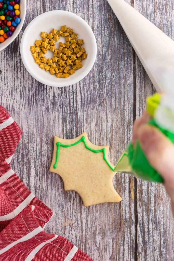 Overhead shot of a Christmas sugar cookie in the shape of a holly leaf.  Green royal icing is being piped around the edge to outline the cookie.