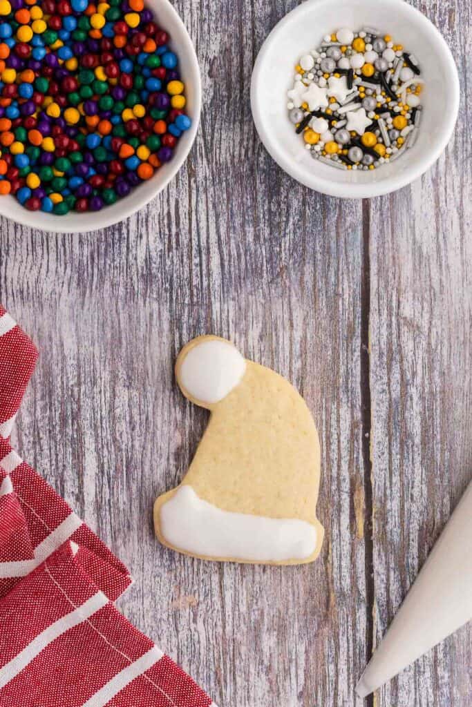 Overhead shot of a Santa hat shaped sugar cookie.  White royal icing has been piped and flooded around the pom pom and down on the bottom trim.  The middle section has been left blank so there is only cookie visible.  