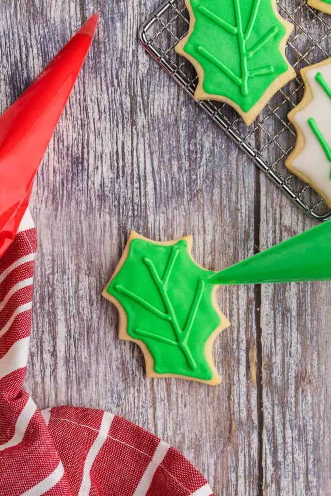 Overhead shot of a sugar cookie shaped like a holly leaf.  It has been piped and flooded with green royal icing and allowed to dry.  Now it is being piped with a thicker green royal icing to show veins in the leaf.