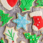 Overhead shot of fully decorated Christmas sugar cookies laying on a wooden counter. Shapes include a Santa's hat which is red with white icing on the pom pom and fringe. The pom pom and fringe have also been covered in white jimmie sprinkles. A blue snowflake with a snowflake design pipe in white. Holly leaves, some green with green piped veins and red holly berries, some white with green piped on veins with red holly berries. A couple of red stockings with white icing at the toe and top fringe. 3 Colored pearl sprinkles have been added to make it look like buttons. A couple of green Christmas trees decorated with medium sized pearl sprinkles and a gold star.