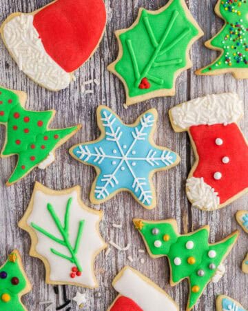 Overhead shot of fully decorated Christmas sugar cookies laying on a wooden counter. Shapes include a Santa's hat which is red with white icing on the pom pom and fringe. The pom pom and fringe have also been covered in white jimmie sprinkles. A blue snowflake with a snowflake design pipe in white. Holly leaves, some green with green piped veins and red holly berries, some white with green piped on veins with red holly berries. A couple of red stockings with white icing at the toe and top fringe. 3 Colored pearl sprinkles have been added to make it look like buttons. A couple of green Christmas trees decorated with medium sized pearl sprinkles and a gold star.