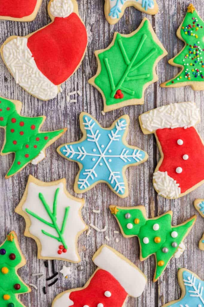 Overhead shot of fully decorated Christmas sugar cookies laying on a wooden counter.  Shapes include a Santa's hat which is red with white icing on the pom pom and fringe.  The pom pom and fringe have also been covered in white jimmie sprinkles.  A blue snowflake with a snowflake design pipe in white.  Holly leaves, some green with green piped veins and  red holly berries, some white with green piped on veins with red holly berries.  A couple of red stockings with white icing at the toe and top fringe.  3 Colored pearl sprinkles have been added to make it look like buttons.  A couple of green Christmas trees decorated with medium sized pearl sprinkles and a gold star.