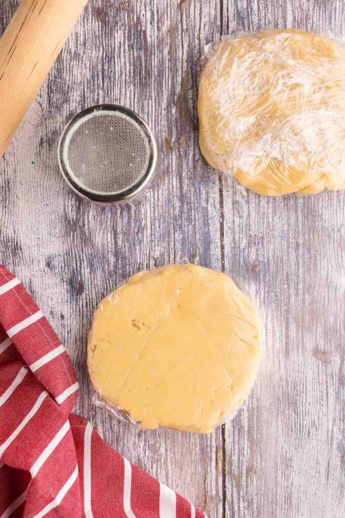 an overhead shot of the christmas sugar cookie dough divided into two discs and wrapped in plastic wrap for chilling.  The discs are about 6 inches in diameter and about 2 inches thick.