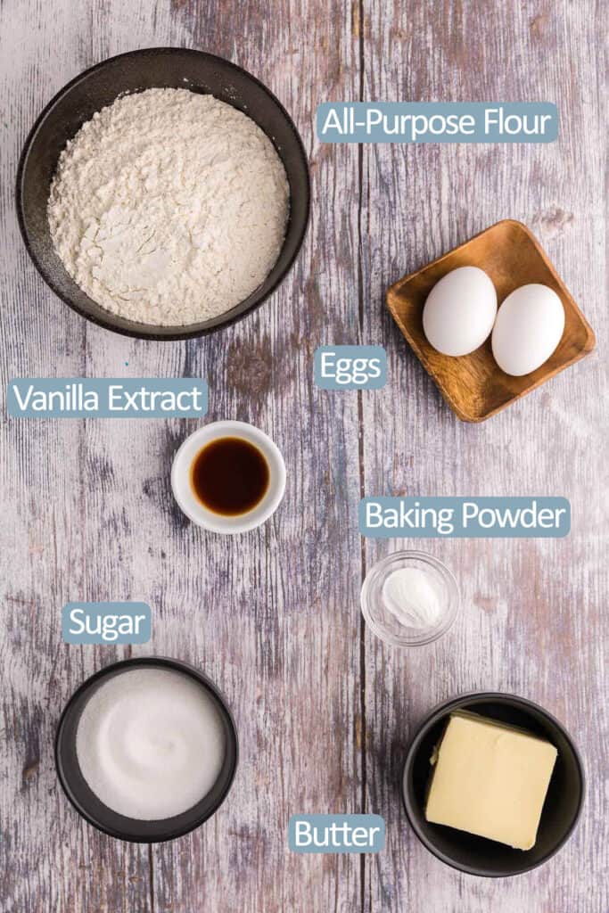 Overhead shot of the ingredients needed to make Christmas Sugar Cookies.  Ingredients include: all-purpose flour, eggs, vanilla extract, baking powder, sugar, and butter.  Not pictured are sprinkles and royal icing if you want to decorate.
