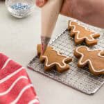 A close up side shot showing a brown gingerbread Christmas tree shaped cookie being decorated with a piping bag of white royal icing. A thin white strip of royal icing has been piped around the entire edge of the tree.
