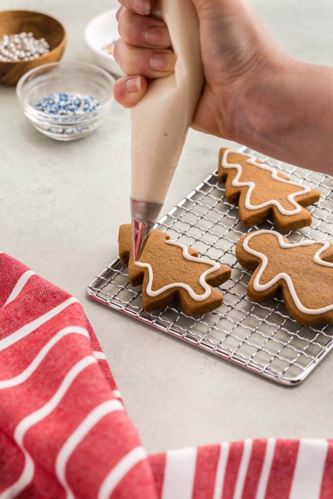 A close up side shot showing a brown gingerbread Christmas tree shaped cookie being decorated with a piping bag of white royal icing. A thin white strip of royal icing has been piped around the entire edge of the tree.