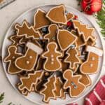 Overhead shot of a round white serving plate piled with gingerbread cookies. The cookies are cut out in the shape of gingerbread women, stockings, christmas trees, and bells. White royal icing has been used to decorate them with an outline and some of them are gently decorated with silver balls, or sprinkles.