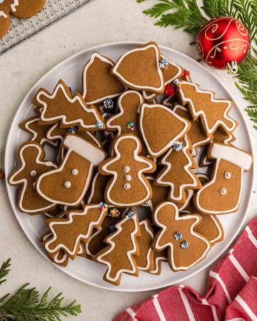 Overhead shot of a round white serving plate piled with gingerbread cookies. The cookies are cut out in the shape of gingerbread women, stockings, christmas trees, and bells. White royal icing has been used to decorate them with an outline and some of them are gently decorated with silver balls, or sprinkles.