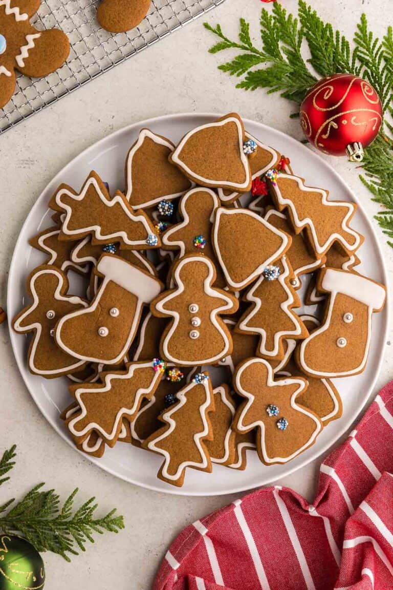 Overhead shot of a round white serving plate piled with gingerbread cookies. The cookies are cut out in the shape of gingerbread women, stockings, christmas trees, and bells. White royal icing has been used to decorate them with an outline and some of them are gently decorated with silver balls, or sprinkles.