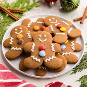 Angled shot of a round white serving plate filled with gingerbread men. Each one has been decorated with royal icing to make two eyes and a smile. The white royal icing also has made squiggly lines on the arms and legs. Each gingerbread man also has two smarties as buttons on its chest. In the background are some cedar boughs, Christmas balls, and cinnamon sticks.