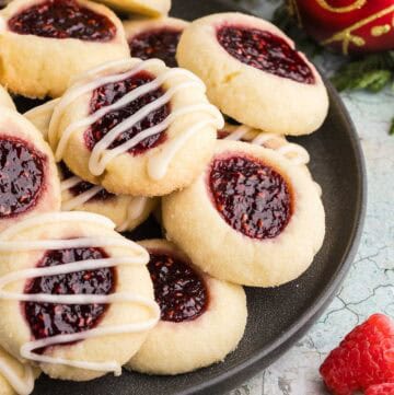 Side shot of a dark grey circular serving plate filled with lightly golden colored raspberry thumbprint cookies. Each cookie top has had an indent filled with ruby-red raspberry jam. Some of the cookies have also been glazed with a few strips of white icing. In the background are some cedar boughs and a red Christmas ball.