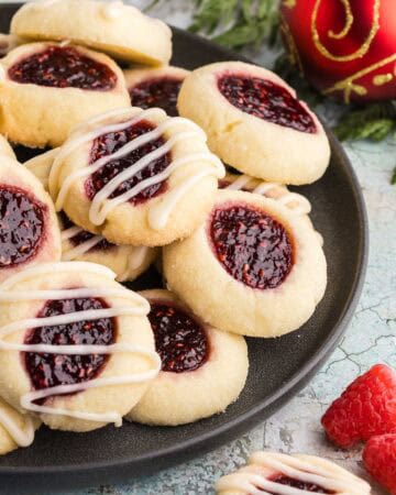 Side shot of a dark grey circular serving plate filled with lightly golden colored raspberry thumbprint cookies. Each cookie top has had an indent filled with ruby-red raspberry jam. Some of the cookies have also been glazed with a few strips of white icing. In the background are some cedar boughs and a red Christmas ball.
