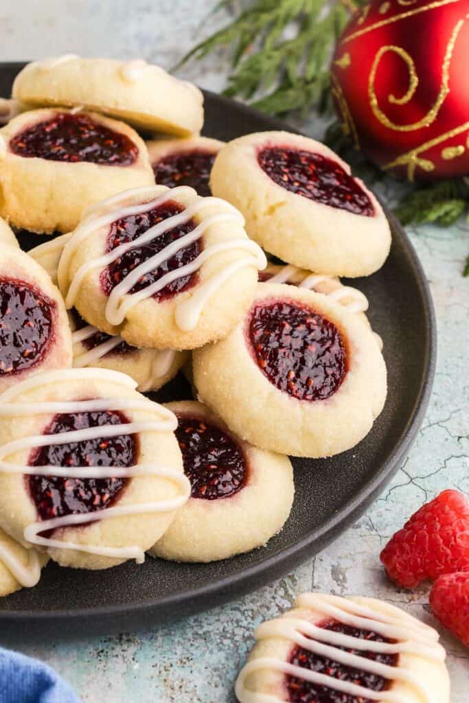 Side shot of a dark grey circular serving plate filled with lightly golden colored raspberry thumbprint cookies. Each cookie top has had an indent filled with ruby-red raspberry jam. Some of the cookies have also been glazed with a few strips of white icing. In the background are some cedar boughs and a red Christmas ball.