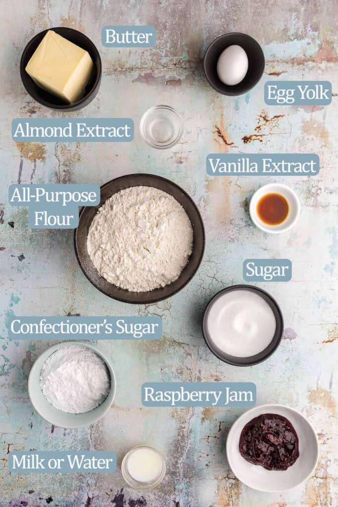 Overhead shot of all the ingredients needed to make raspberry thumbprint cookies. Ingredients are: butter, egg yolk, almond extract, vanilla extract, all-purpose flour, sugar, confectioner's sugar, milk or water, raspberry jam.