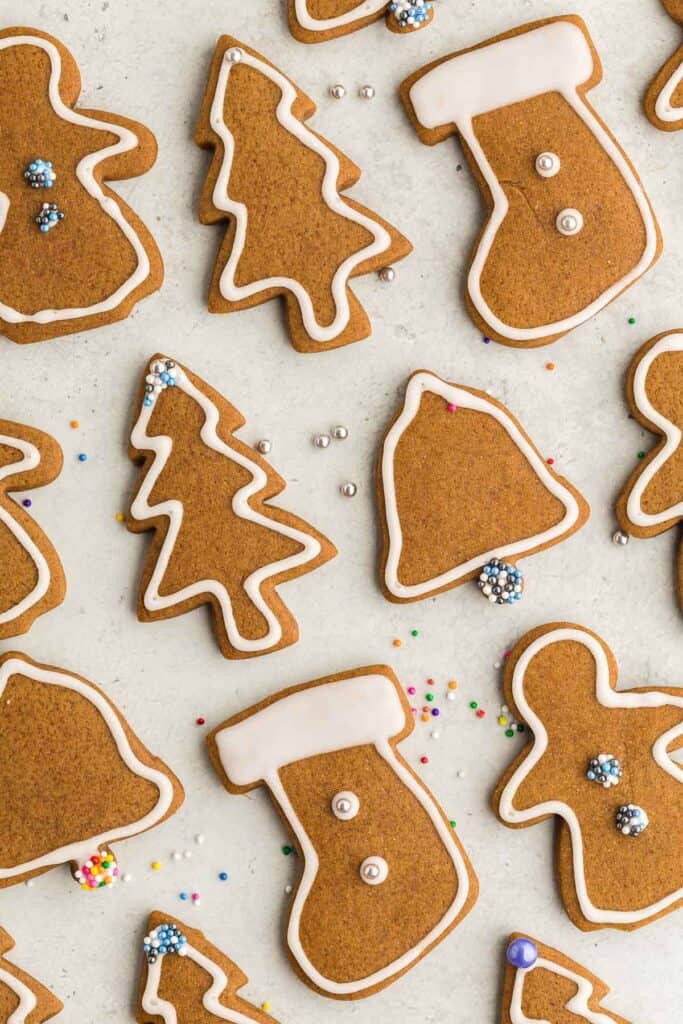 Overhead close up of gingerbread cookies decorated with royal icing and some with sprinkles. The royal icing has been piped around the edge of each cookie. Some are Christmas trees, gingerbread men, bells, and stockings. The stockings have had the top of the stocking flooded with white royal icing to make it seem like a fuzzy white top.
