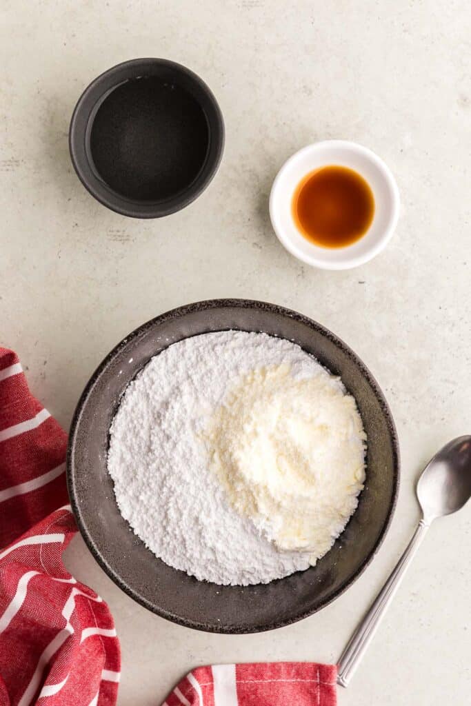 Overhead shot of a black ceramic bowl filled with white confectioners sugar and on top is a small pile of off white meringue powder. Above the black bowl are two pinch bowls. One filled with dark vanilla extract and the other with water. A spoon is lying beside the bowl which will be used to mix everything together.