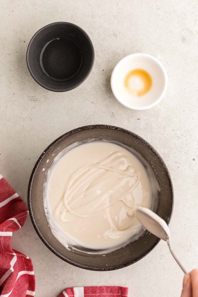 Overhead shot of a black ceramic bowl filled with royal icing. A spoon is drizzling the icing back in to the bowl to show the consistency of the icing. Just soft lines are present.