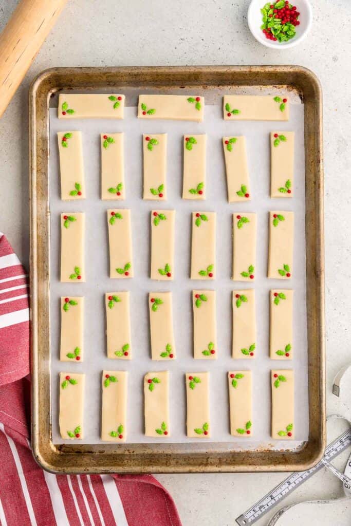 An overhead shot of a parchment lined baking sheet filled with 27 rectangular shortbread cookies.  Each cookie has been decorated in it's lower left corner and upper right corner with two green holly leaves candy and a red candy berry.